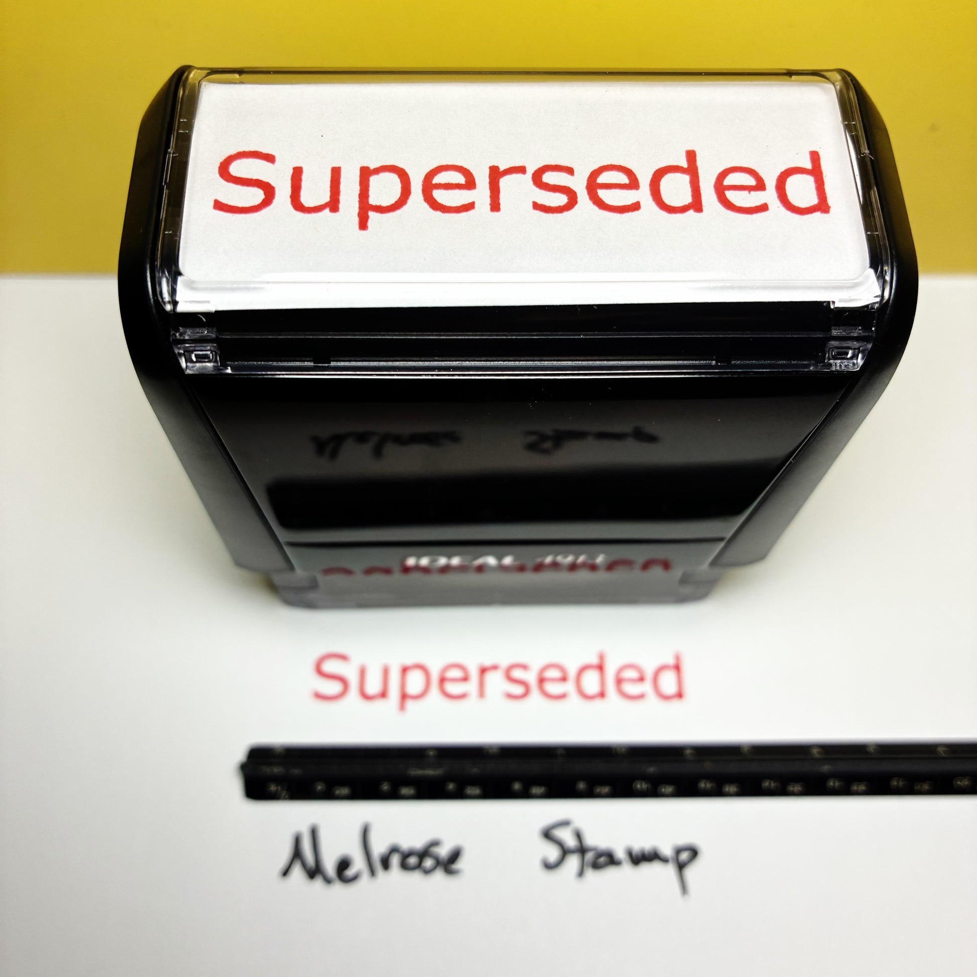 Rubber stamp with 'Superseded' text red ink on a white surface, next to a ruler for scale.