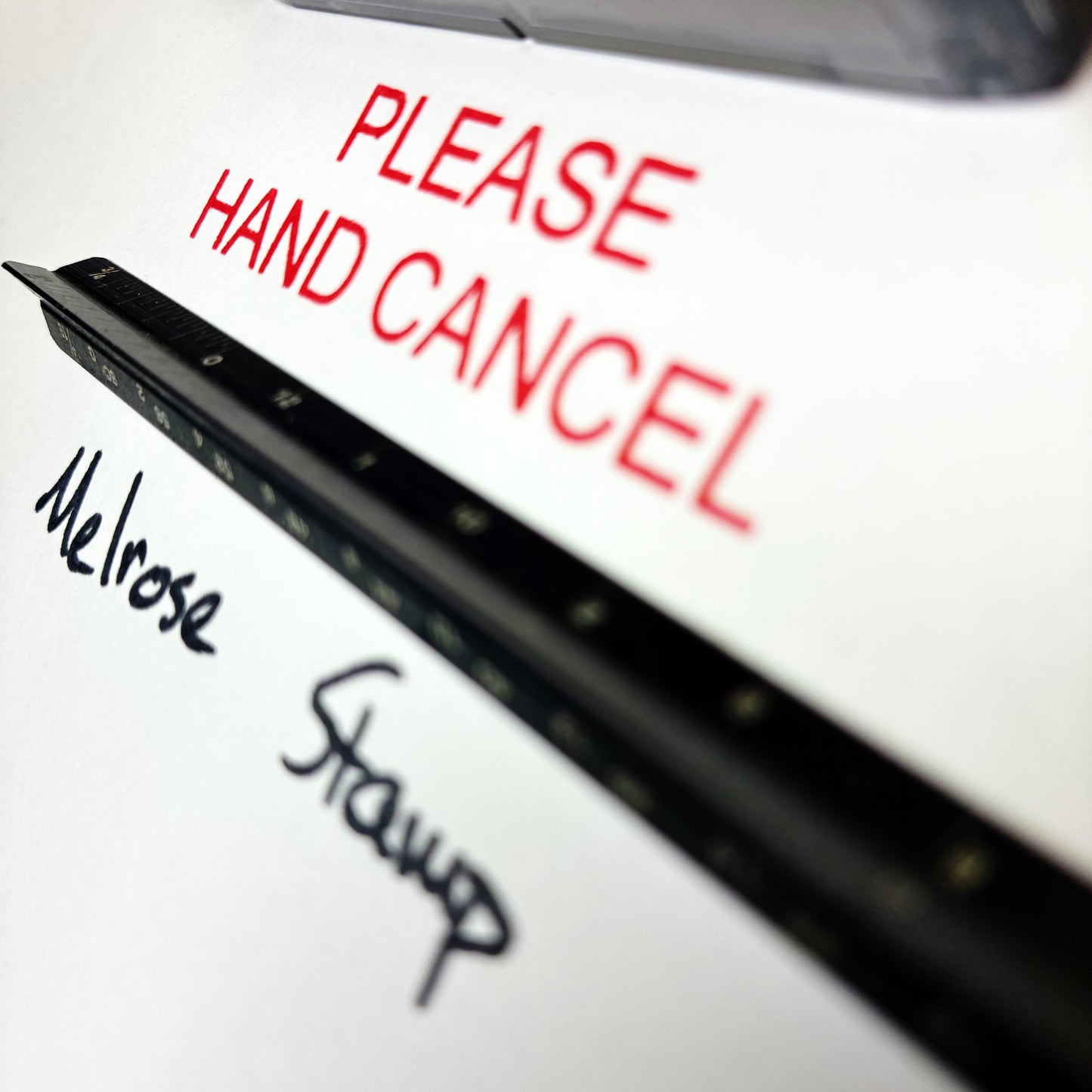 Close-up of a sign with 'PLEASE HAND CANCEL' in red text, with 'Melrose Stamp' written below. Rubber stamp red ink