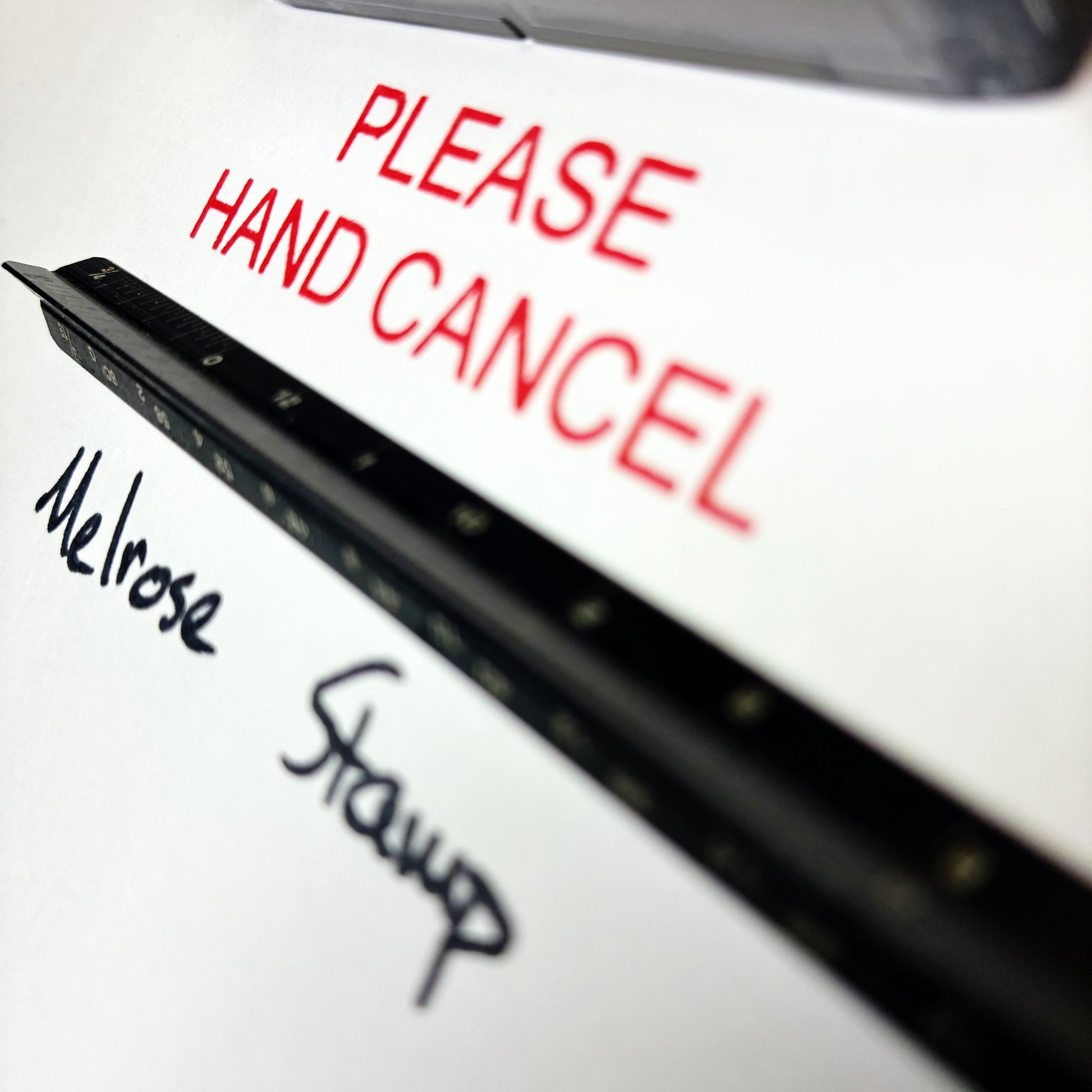 Close-up of a sign with 'PLEASE HAND CANCEL' in red text, with 'Melrose Stamp' written below. Rubber stamp red ink
