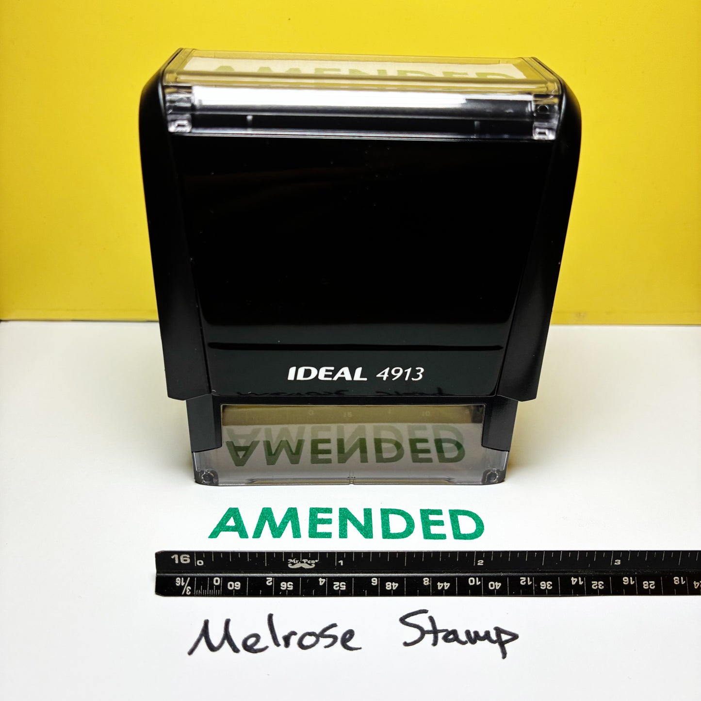 Rubber stamp with 'AMENDED' green ink self-inking on a white surface and yellow background