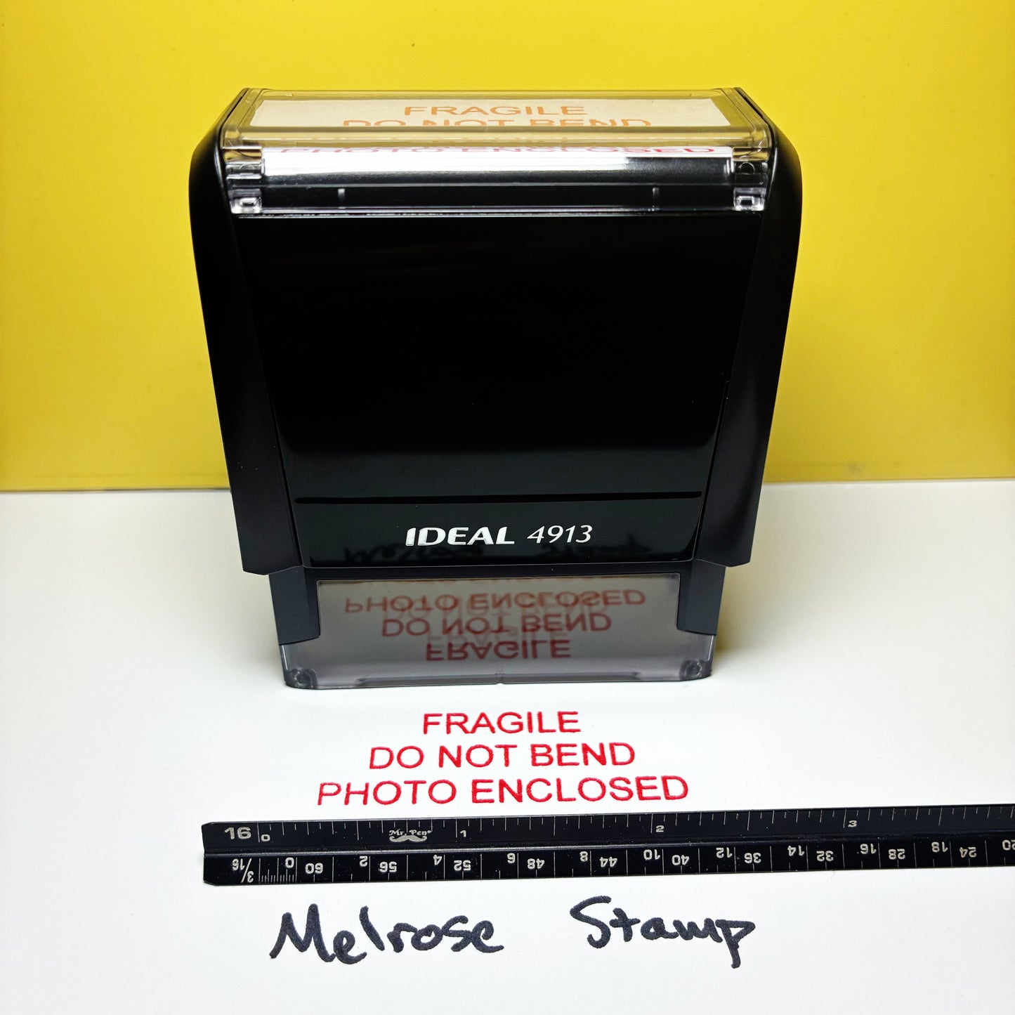 Rubber stamp with 'Fragile Do Not Bend Photo Enclosed' text on a white surface with a yellow background.