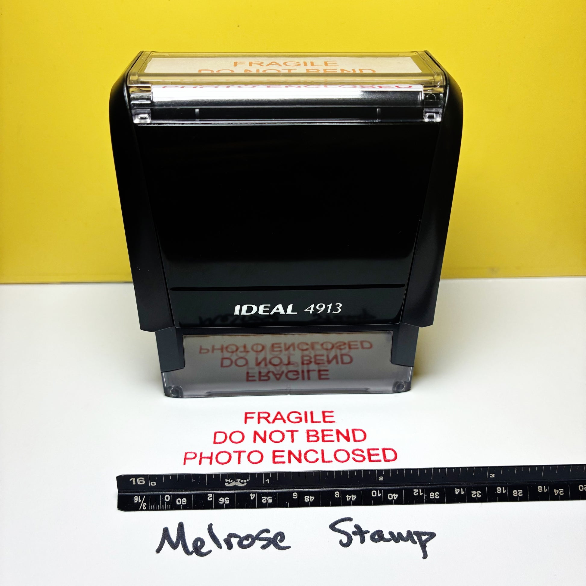 Rubber stamp with 'Fragile Do Not Bend Photo Enclosed' text on a white surface with a yellow background.