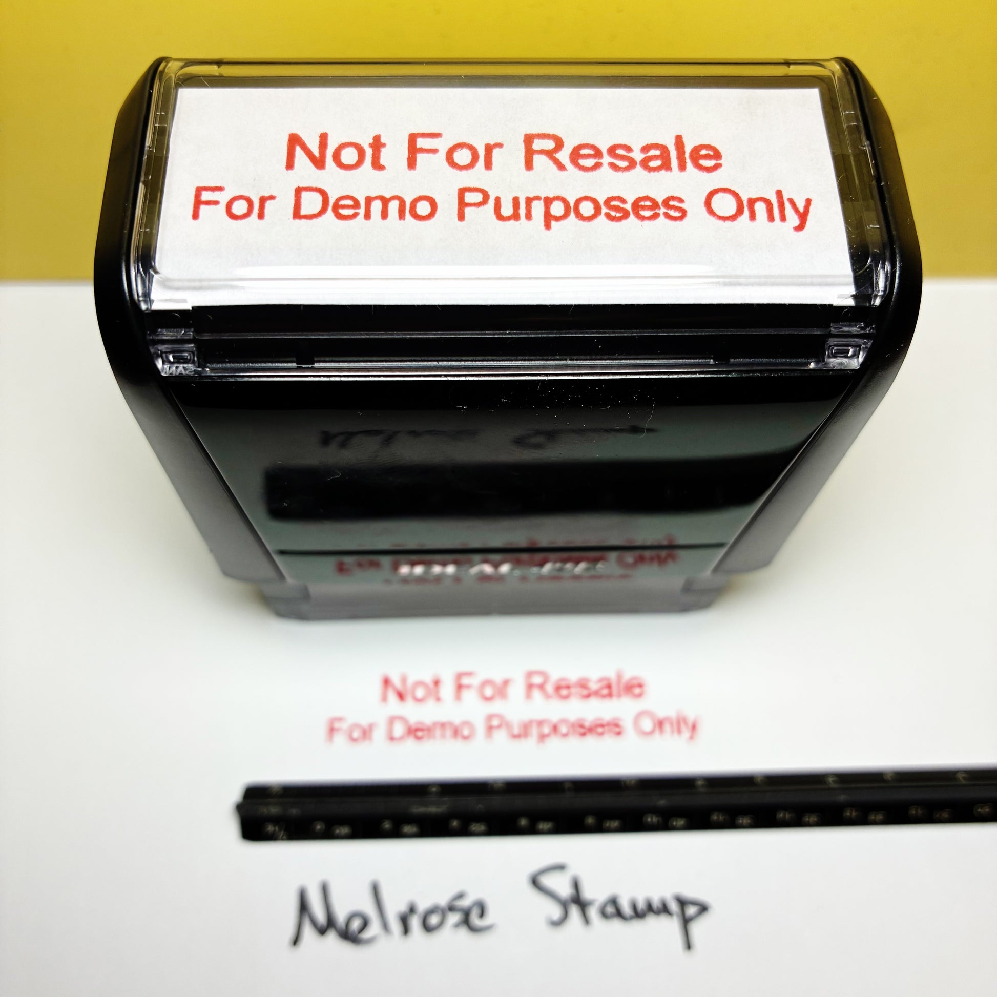 A black self-inking stamp with a white paper insert that reads 'Not For Resale For Demo Purposes Only' in red text. The stamp is shown on a white surface against a yellow background, with a black ruler below and the words 'Melrose Stamp' written underneath.