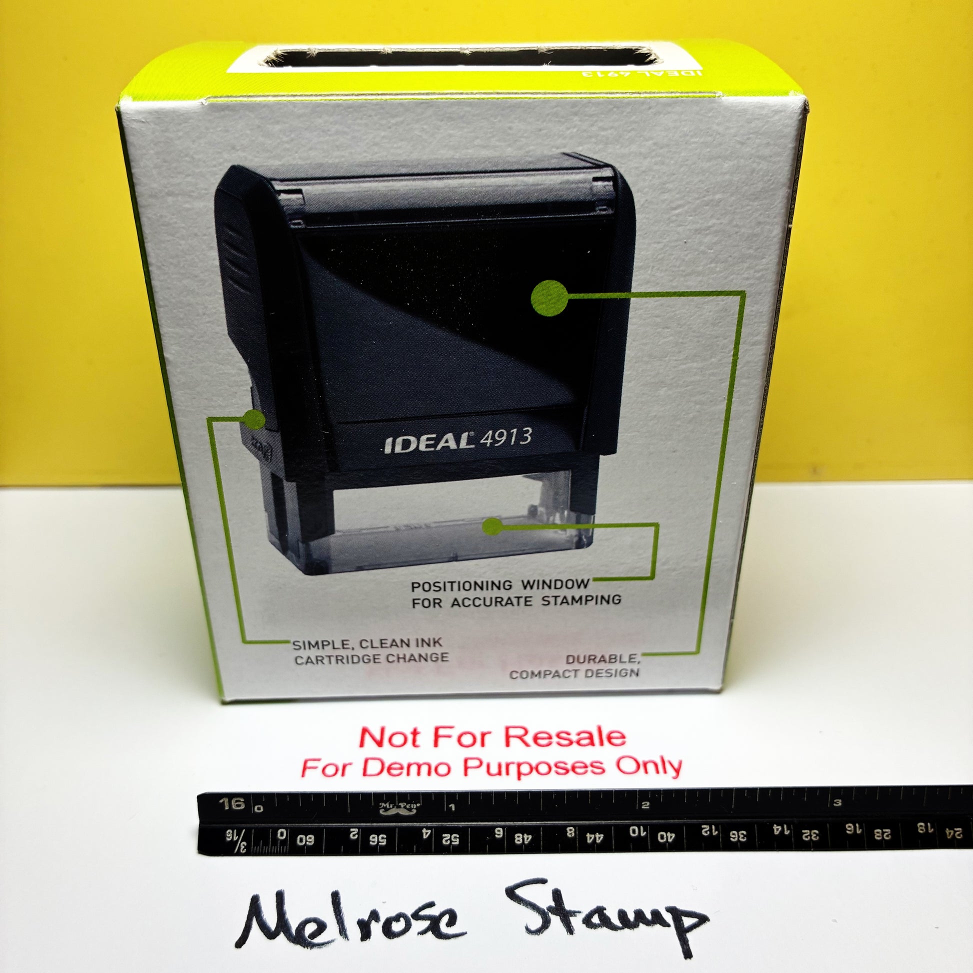 A black Ideal 4913 desktop stamp shown in front of its white retail box with green line graphics, placed on a white surface against a yellow background. A black ruler and handwritten text reading 'Melrose Stamp' are visible below.