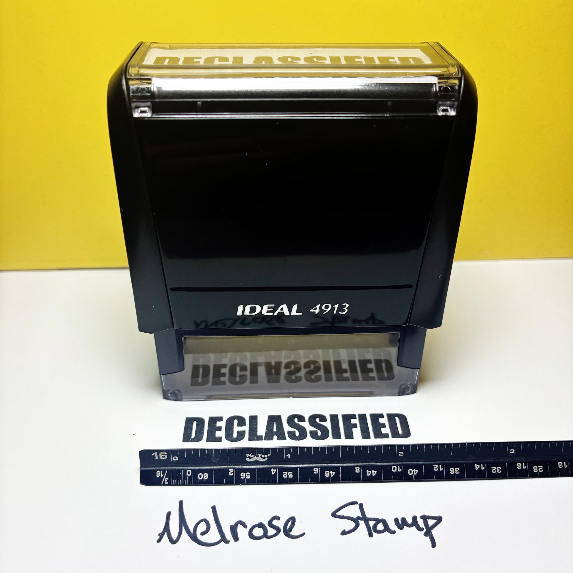 Black stamp labeled 'DECLASSIFIED' with a ruler for scale on a white surface against a yellow background.