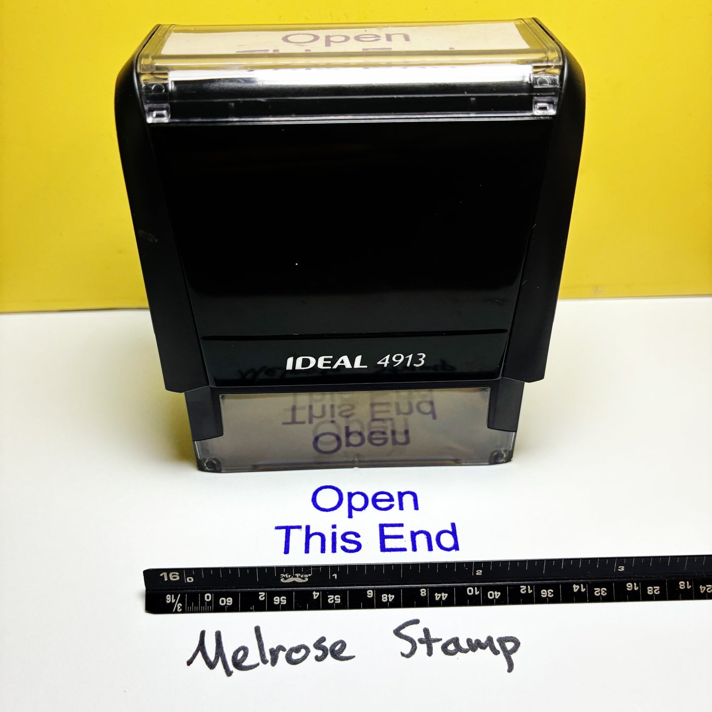 Black stamp with 'Open This End' text and ruler for scale on a yellow and white background
