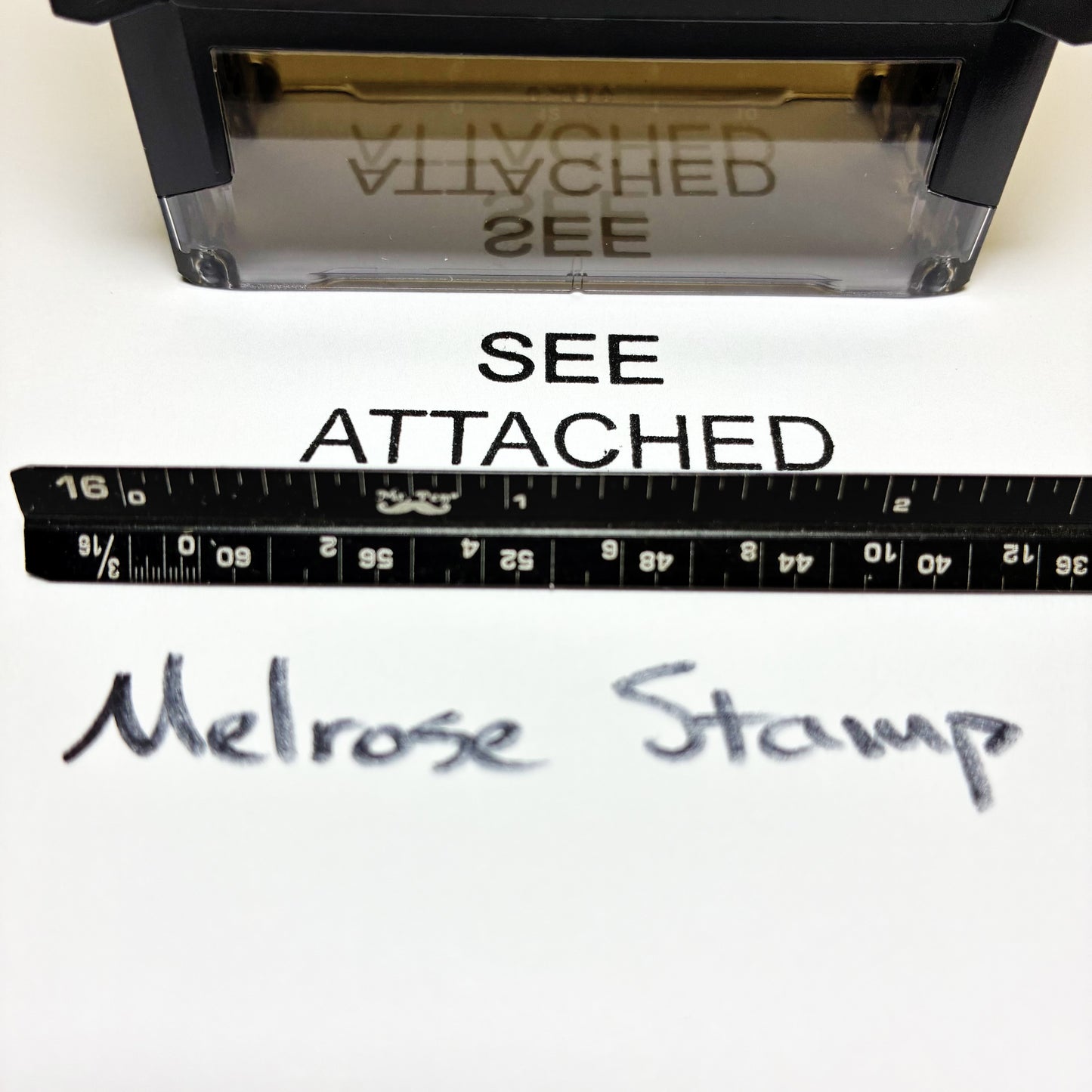 Rubber stamp with text 'SEE ATTACHED' and 'Melrose Stamp' next to a ruler on a white background.